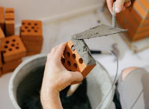 One young unrecognizable Caucasian man applying fresh cement mortar to a brick using a trowel for the chimney of a fireplace in a room, close-up side view with selective focus. Clean brickwork concept, home renovation.